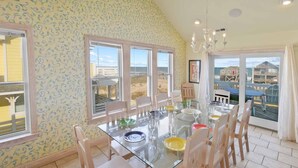 Un comedor con mesa y sillas de vidrio, ventanas amplias con vista a la playa y una lámpara de araña.