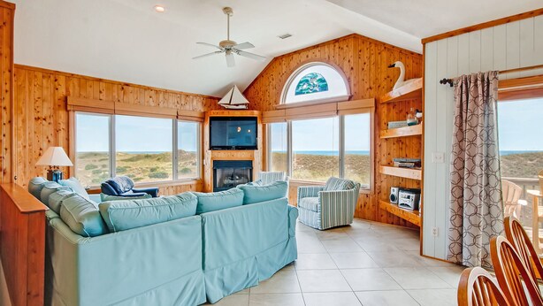 A spacious living room with a large blue sofa, a fireplace, a television, and a view of the desert landscape through large windows. - Oceanfront Home in Waves w/ Private Pool, Hot Tub, Game Room. Total Seaduction. (Waves)