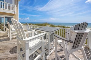 A deck with white Adirondack chairs and a table, overlooking a beach with the ocean in the background.