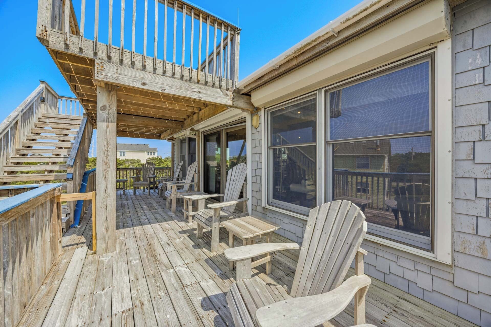 A wooden deck with Adirondack chairs and a small table, a covered area with a railing, and a large window on the side of a building.