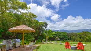 Un espace barbecue équipé de grils, d’un parasol en chaume et de chaises rouges, aménagé dans un cadre extérieur verdoyant, avec des arbres et des montagnes en arrière-plan.