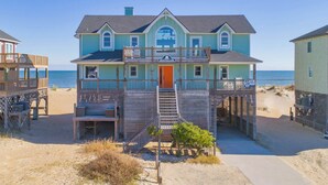 Een huis aan het strand met een balkon en een trap naar de ingang, met een bord dat surflessen aanbiedt.
