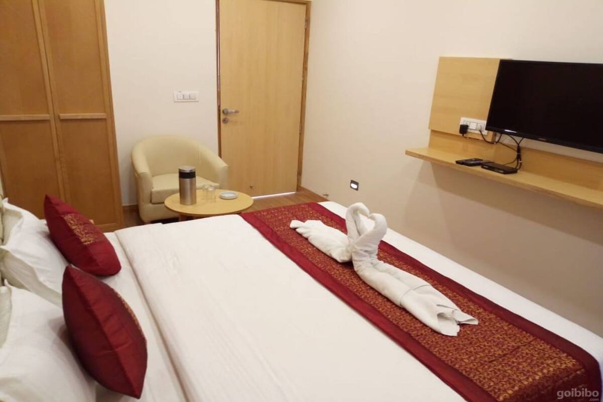 A hotel room with a neatly made bed, a chair, a small table with a water bottle, and a TV on a shelf.