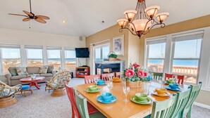 A spacious living room with a wooden dining table set for six, surrounded by colorful chairs. A large window offers a view of the beach. - Semi-Oceanfront Dog Friendly w/ Private Pool, Hot Tub, Game and Media Rooms (Rodanthe)
