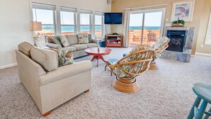 A spacious living room with a sofa, armchair, and a small table. There is a television mounted on the wall and a fireplace. - Semi-Oceanfront Dog Friendly w/ Private Pool, Hot Tub, Game and Media Rooms (Rodanthe)