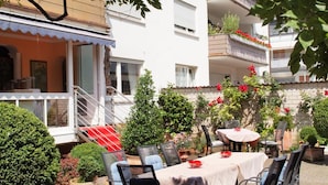 An outdoor dining area with a table and chairs, surrounded by potted plants and a staircase leading to an upper floor.