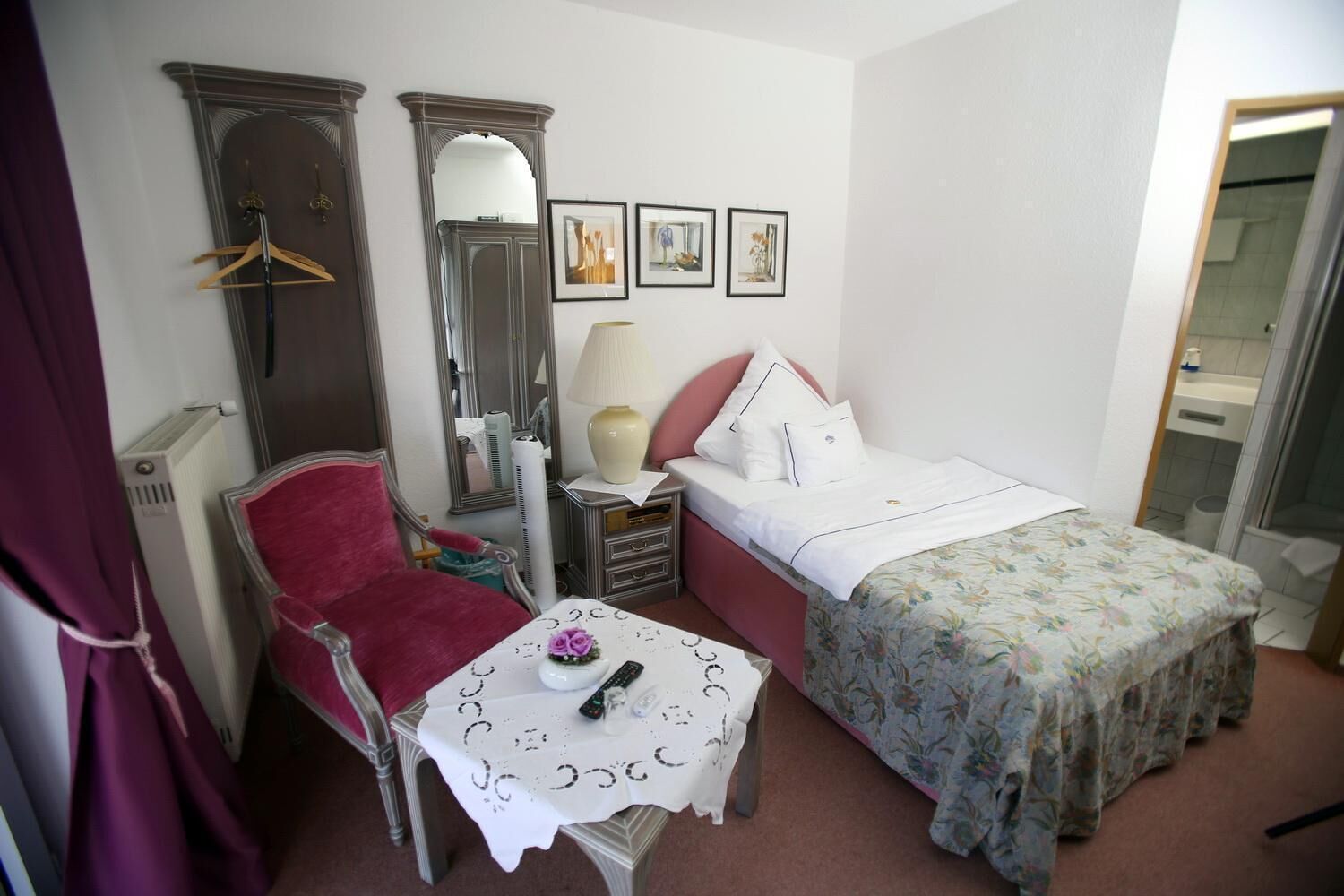 A small, compact bedroom with a single bed, a chair, a table with a vase, and a mirror.