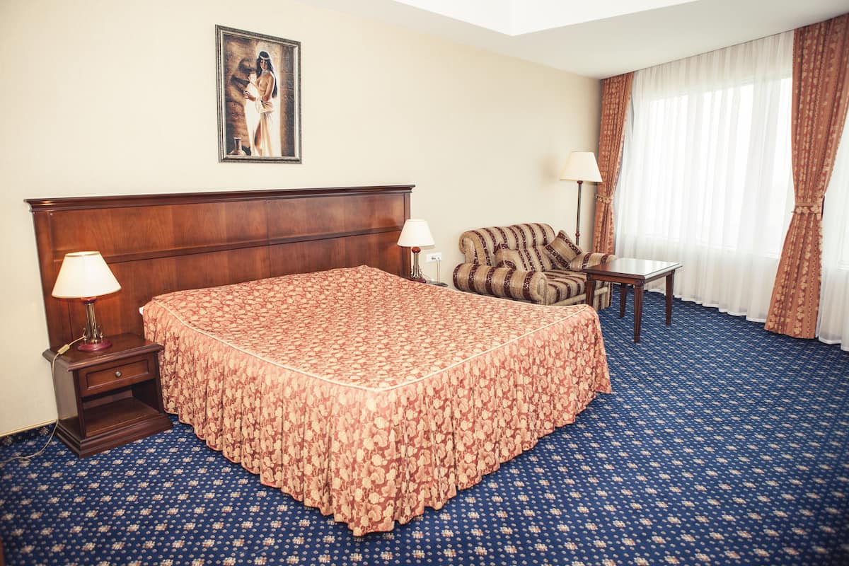 A hotel room with a large bed, a chair, a small table, a lamp, and a painting on the wall.