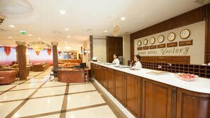 A hotel reception area with a check-in counter, a seating area with sofas, and a wall displaying clocks and city names. - Grand Hotel Victory (Aktau)