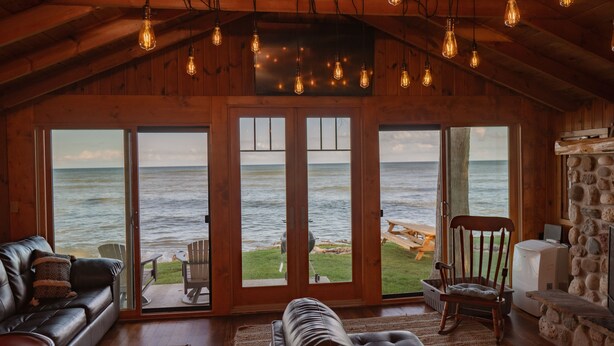 Una cabaña acogedora con un amplio ventanal corredera que da a un patio con vistas al océano. En el interior, hay un sofá de cuero, un sillón mecedor y una chimenea de piedra.