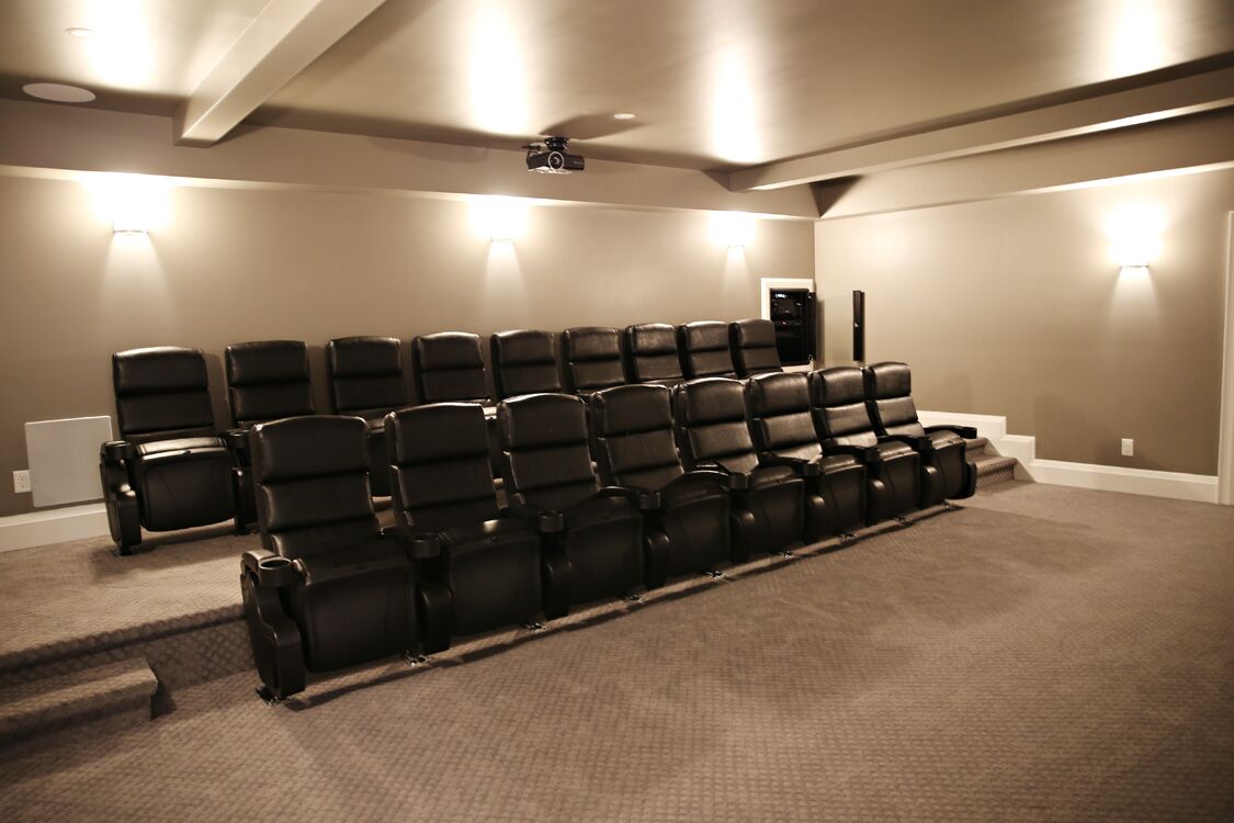 A home theater room with rows of black leather chairs, carpeted floor, and a ceiling-mounted projector.