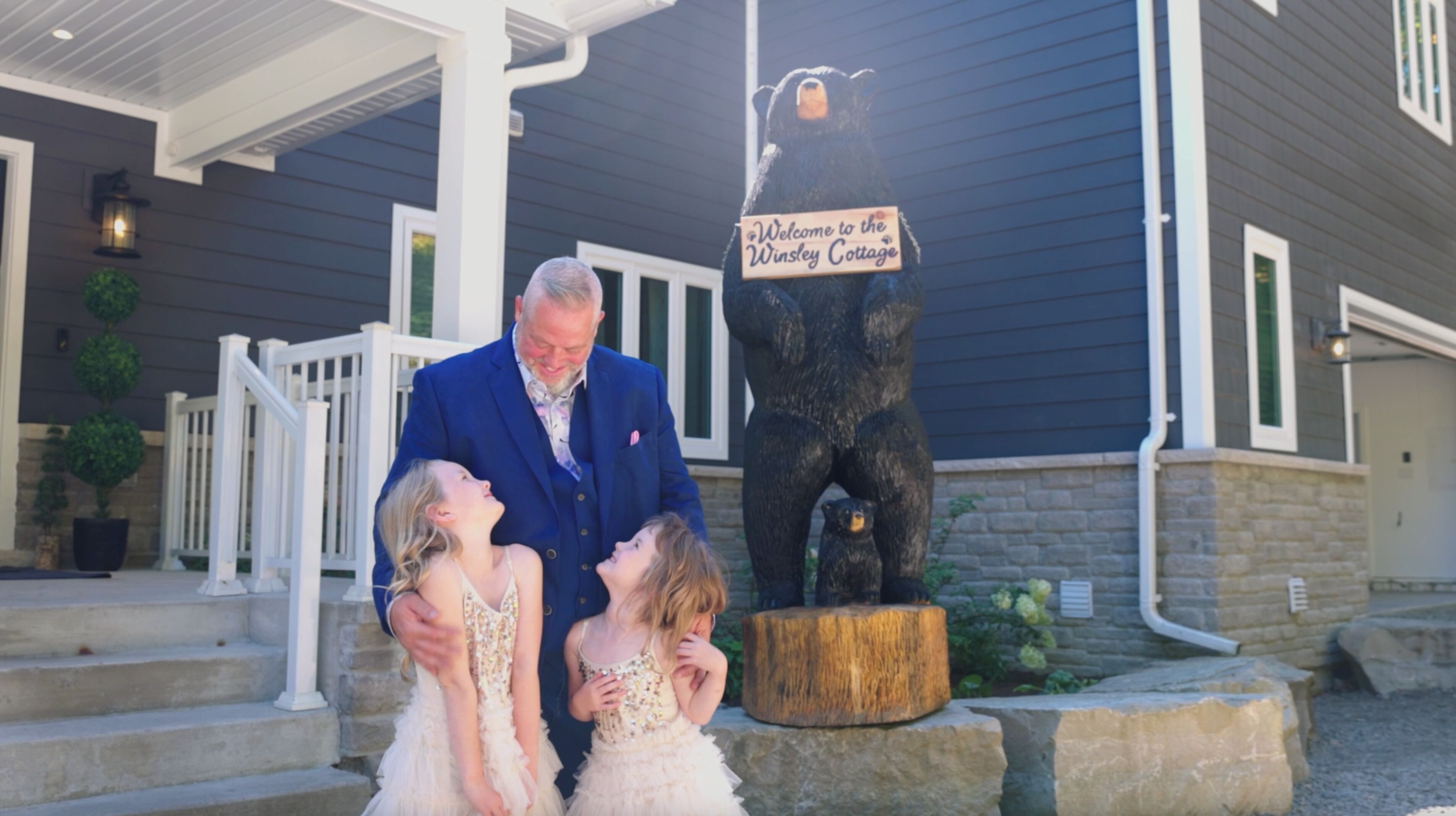 Un hombre y dos niños frente a una casa con una estatua de madera de un oso y un cartel que dice 'Welcome to the Winstley Cottages'.
