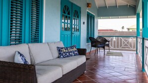 A covered outdoor seating area with a sofa and chairs, turquoise shutters, and a view of the surrounding area. - Lavaughn House - Restorations Tobago (Bon Accord)