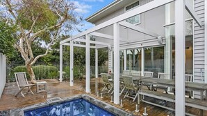 Zona de piscina exterior moderna con terraza de madera, pérgola blanca y asientos con cerramiento de vidrio.
