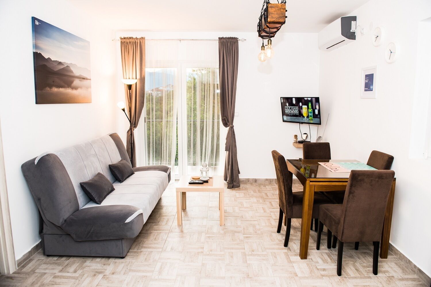A modern living room with a sofa, dining table, and chairs. There is a TV on the wall and a painting above the sofa.