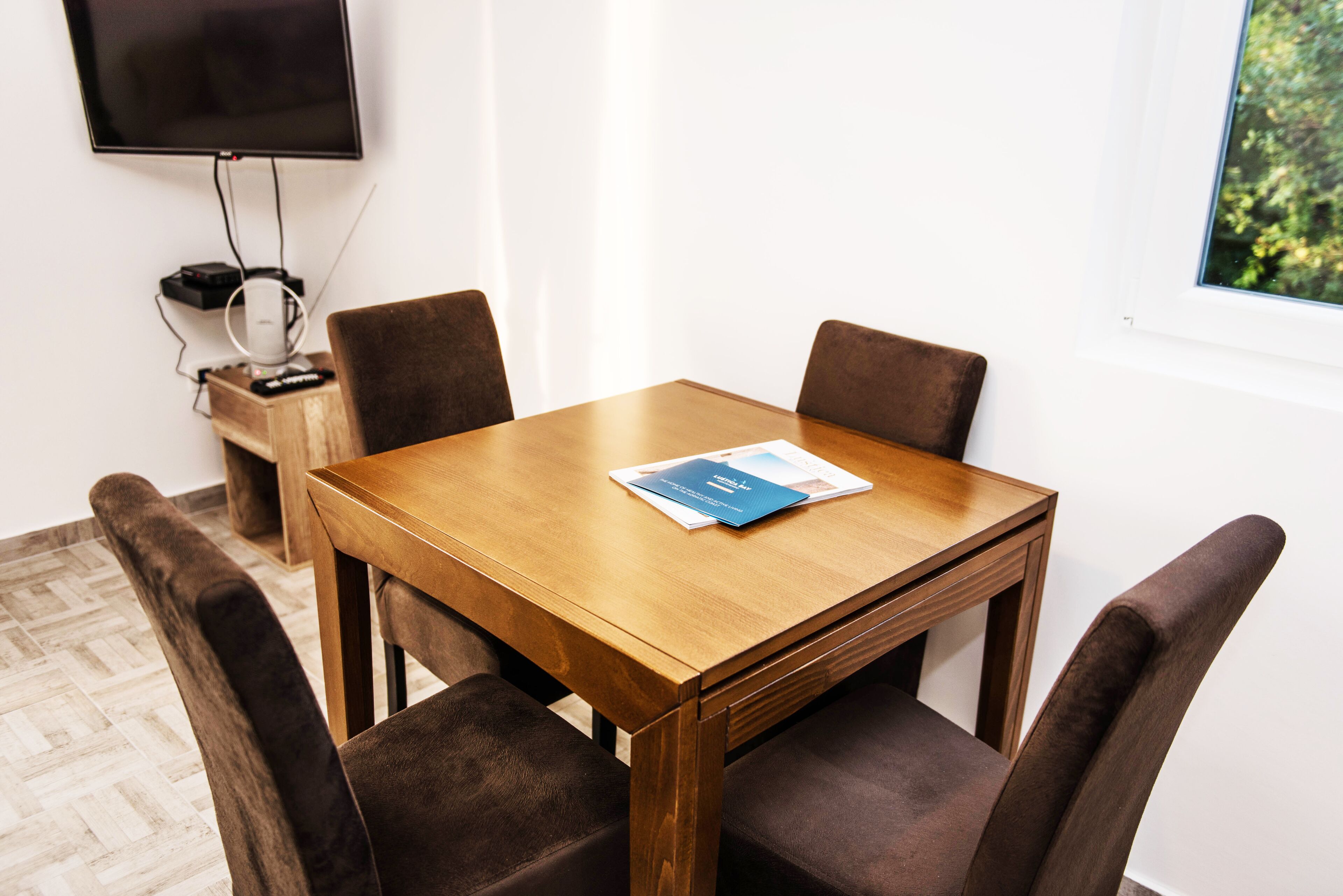 Uma sala de conferências com uma mesa de madeira, quatro cadeiras, uma TV de tela plana e uma janela com vista para o verde.
