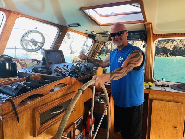 A man steering a boat with a wooden interior, steering wheel, and various controls.