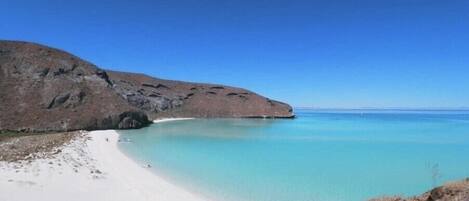A beach with clear turquoise water, white sand, and rocky hills.