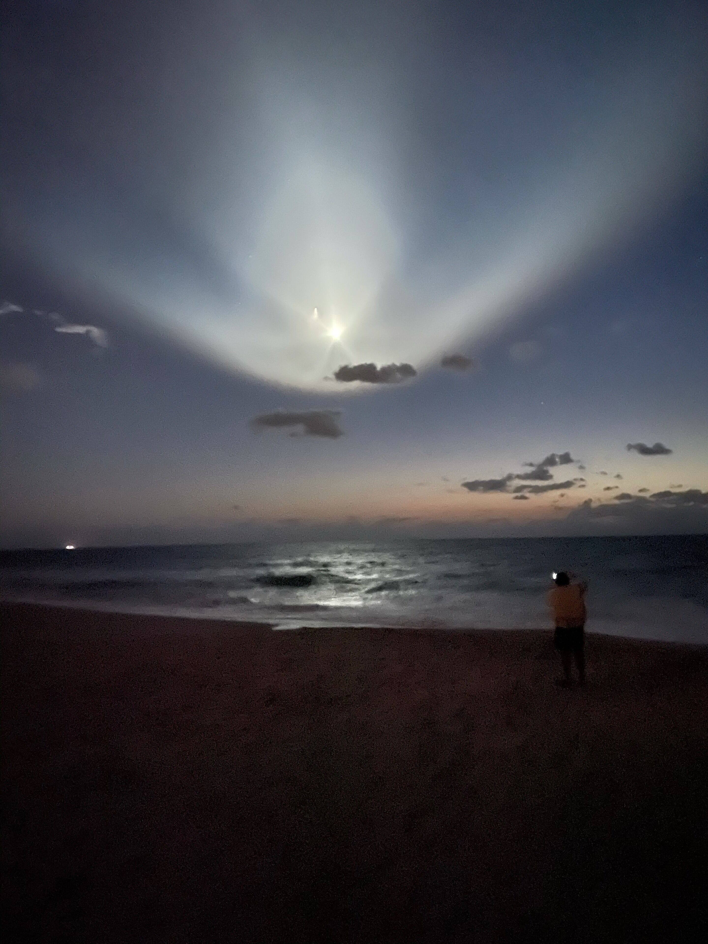 Une scène de plage où l’on voit une personne debout, face à l’océan, avec une lune visible et une source de lumière lointaine.