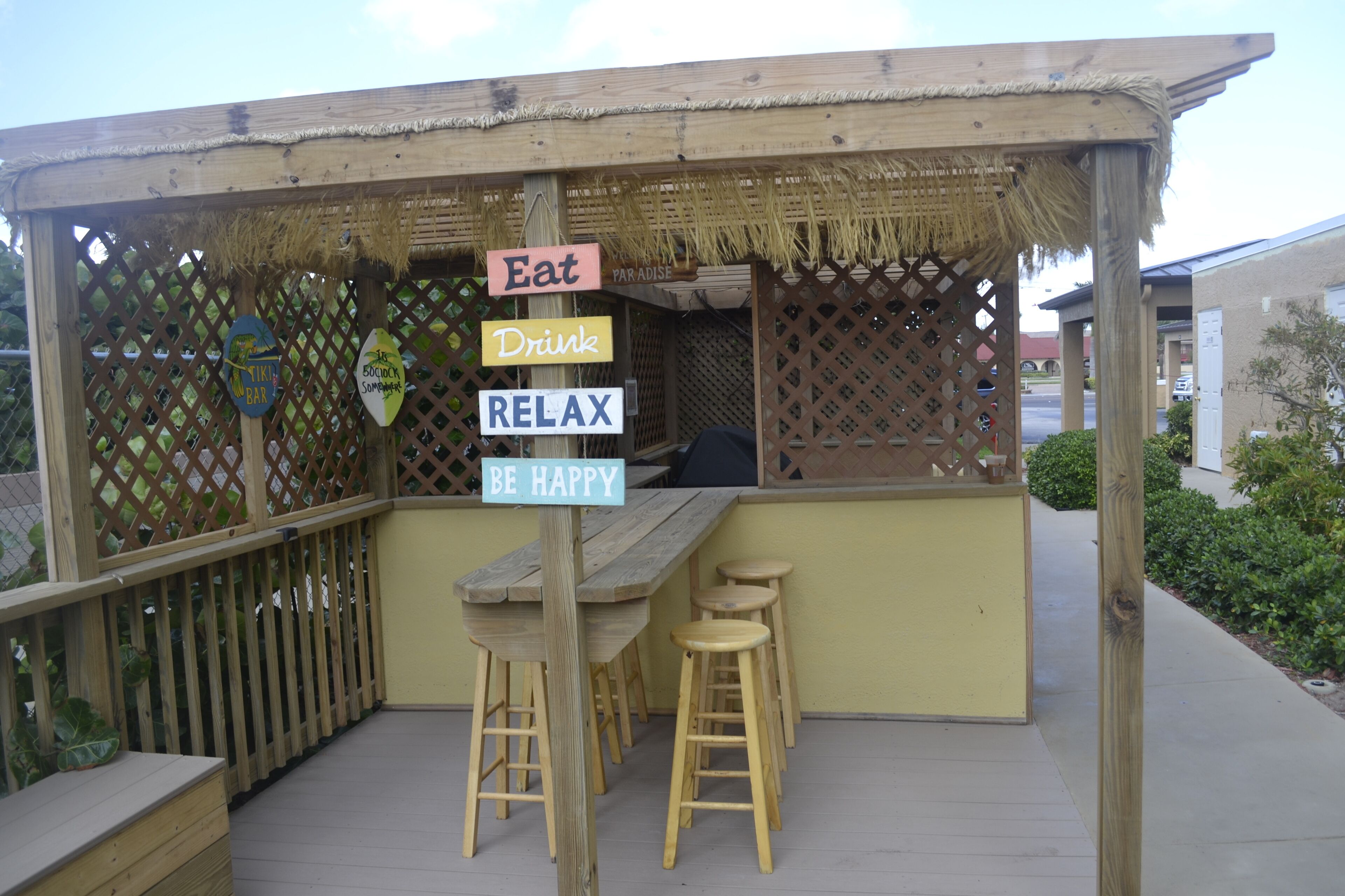 A wooden outdoor seating area with a thatched roof, a wooden table, and bar stools.
