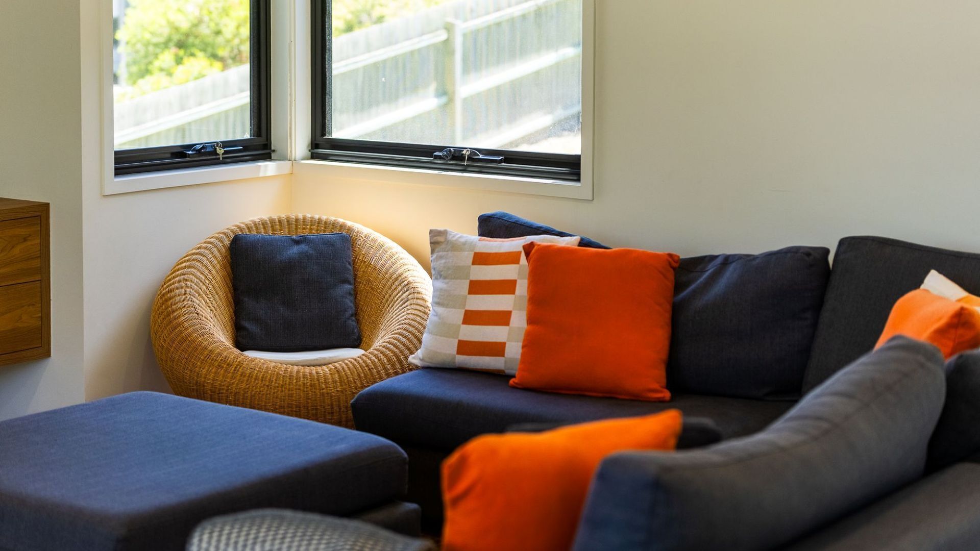 A cozy living area with a wicker chair, a sofa with orange and blue cushions, and a window with a view of a fence.