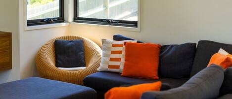 A cozy living area with a wicker chair, a sofa with orange and blue cushions, and a window with a view of a fence.