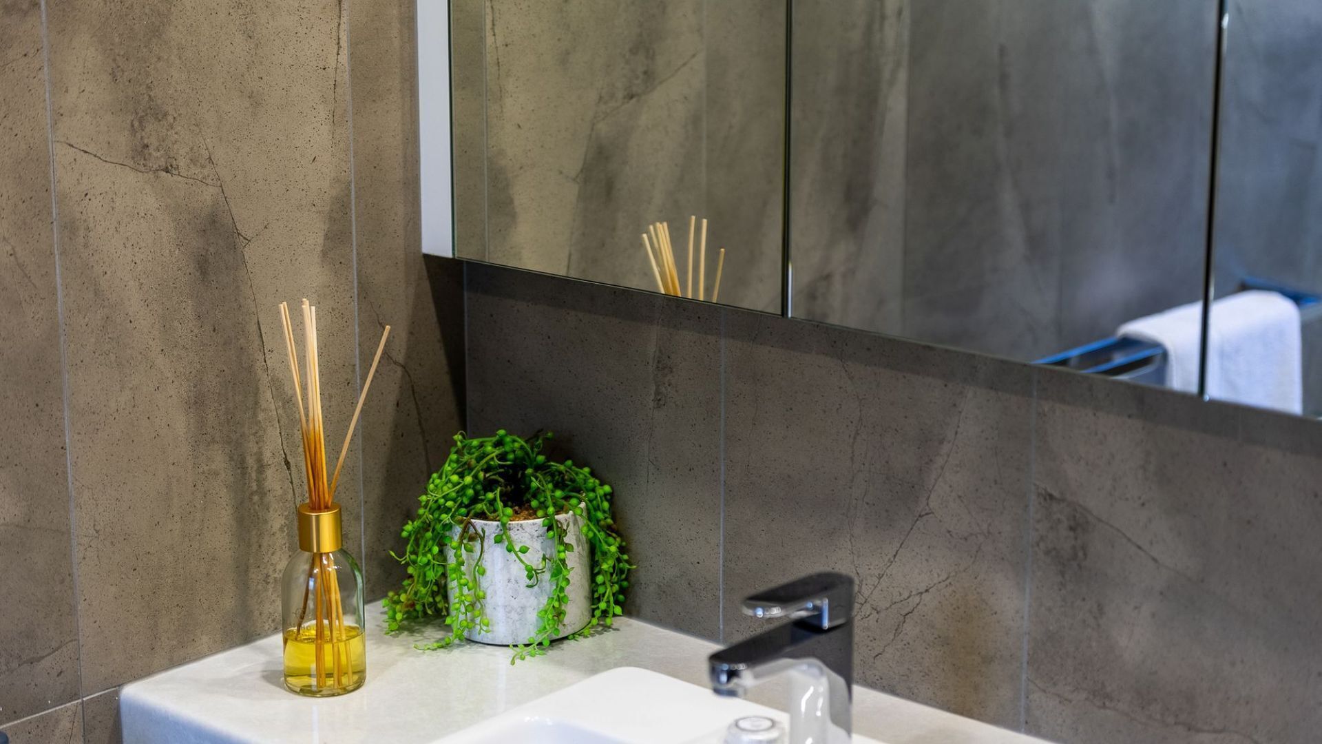 A bathroom with a white sink, a mirror, a bottle of liquid, reed diffusers, and a small potted plant.