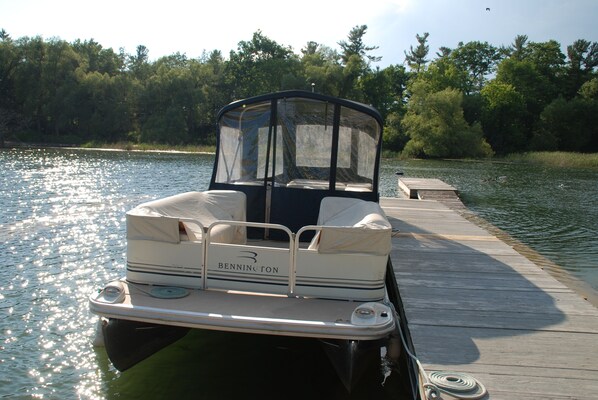 A pontoon boat docked at a wooden pier with a canopy, surrounded by trees.