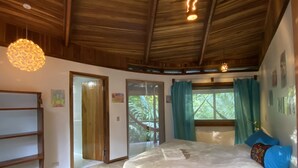 A bedroom with a wooden ceiling, a bed with a white mattress, a wooden bench, and a door leading outside.