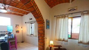 Una habitación con suelo de baldosas, un pilar de ladrillo, un banco de madera, una mesa con lámpara, una ventana con cortinas y una zona de cocina con fregadero y sillas.