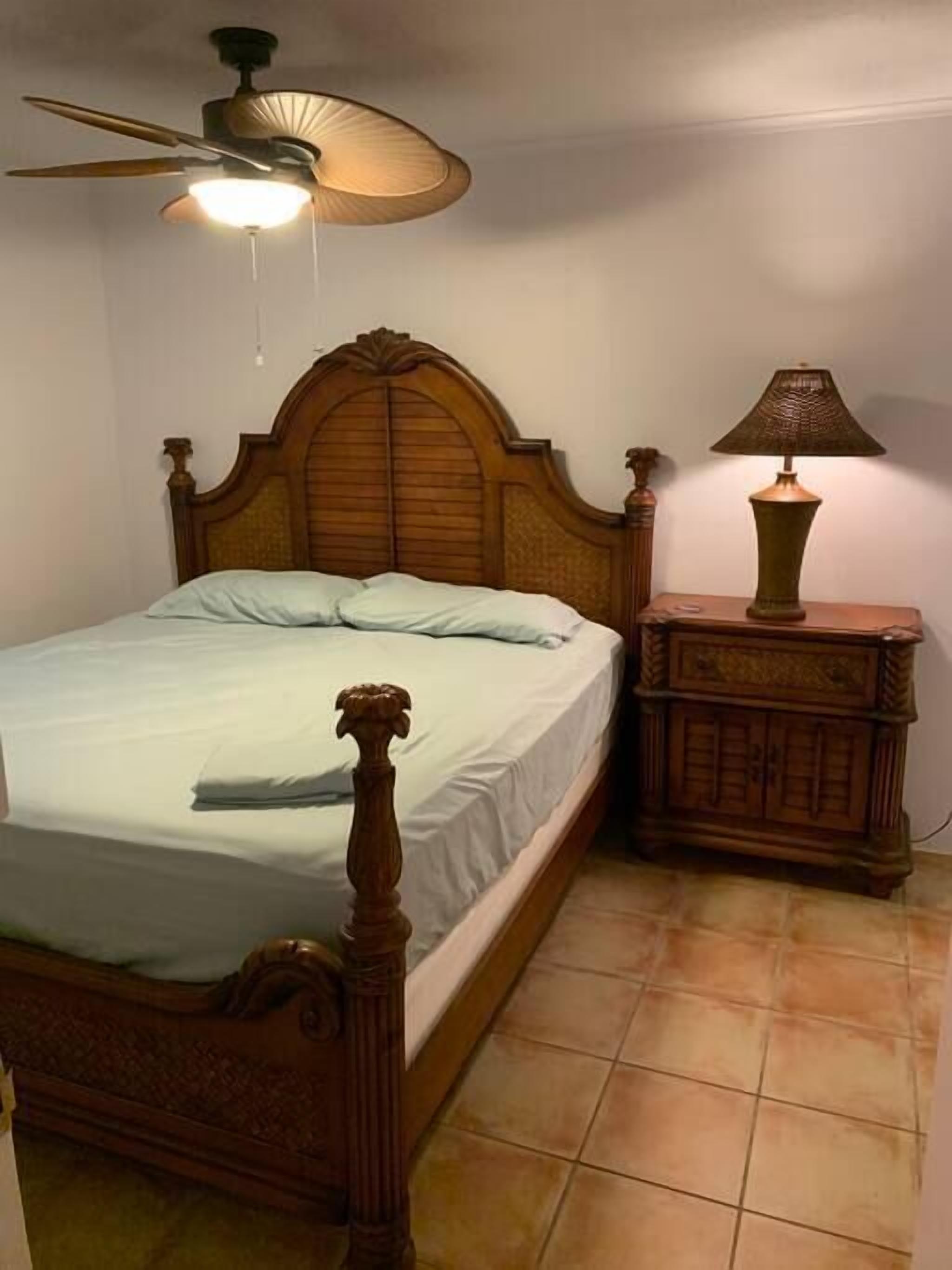 Une chambre à coucher avec un lit en bois, une table de chevet, une lampe et un ventilateur de plafond.