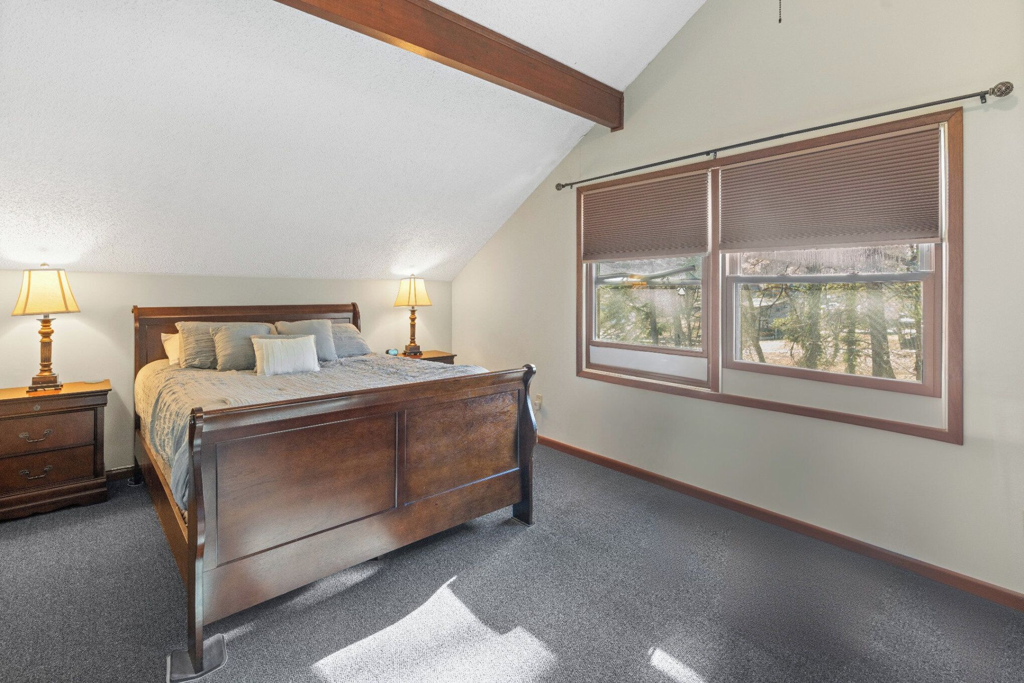 Une chambre à coucher avec un lit en bois, deux tables de chevet avec des lampes, une fenêtre avec des stores et une vue sur les arbres à l’extérieur.