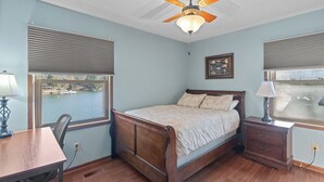 Quarto com cama de madeira, escrivaninha e cadeira. Há uma janela com persianas, um ventilador de teto e mesa de cabeceira com luminária.
