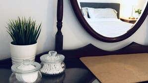 Ein ordentlich eingerichtetes Hotelzimmer mit einem Bett, einem Spiegel, einer Topfpflanze und einem kleinen Tisch mit Taschentuchbox und einem Einmachglas.