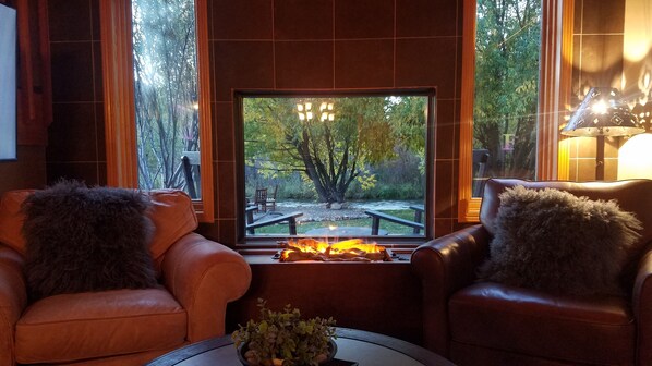 Ein gemütlicher Sitzbereich mit Ledersesseln und Kamin, mit Blick auf einen Garten mit Bäumen und einer Holzterrasse.