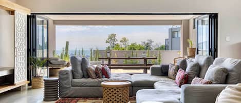 A modern living room with a grey sofa set, a patterned rug, and a view of greenery and buildings through large windows.