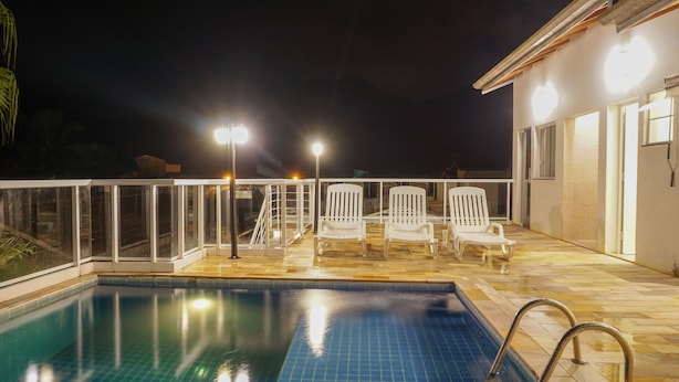 Piscina com deck revestido, cercada por guarda-corpo de vidro e três cadeiras de sol brancas.