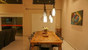 Uma área de refeições com uma mesa de madeira, cadeiras e luminárias pendentes. Há uma pintura na parede e um sofá na sala adjacente.
