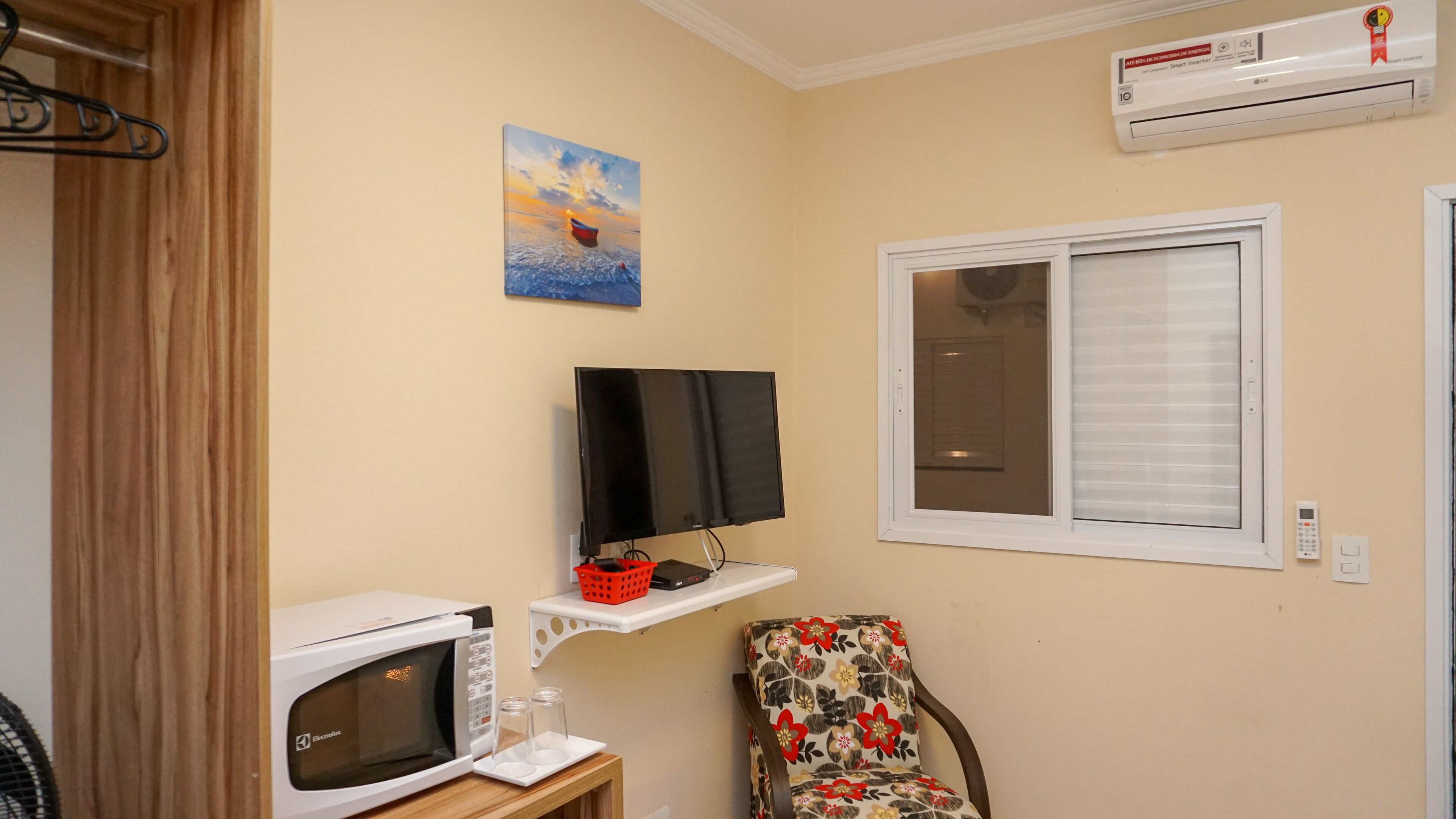 A compact room with a microwave, a flat-screen TV, a window with blinds, a chair, and a painting on the wall.