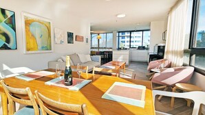 Un comedor con una mesa de madera, sillas y una botella de champán. Hay obras de arte en las paredes y se ve la cocina y el salón al fondo.