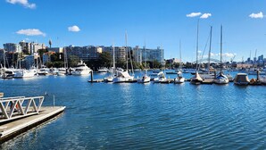 Un puerto deportivo con varias embarcaciones amarradas, un horizonte urbano al fondo y un cielo azul despejado.