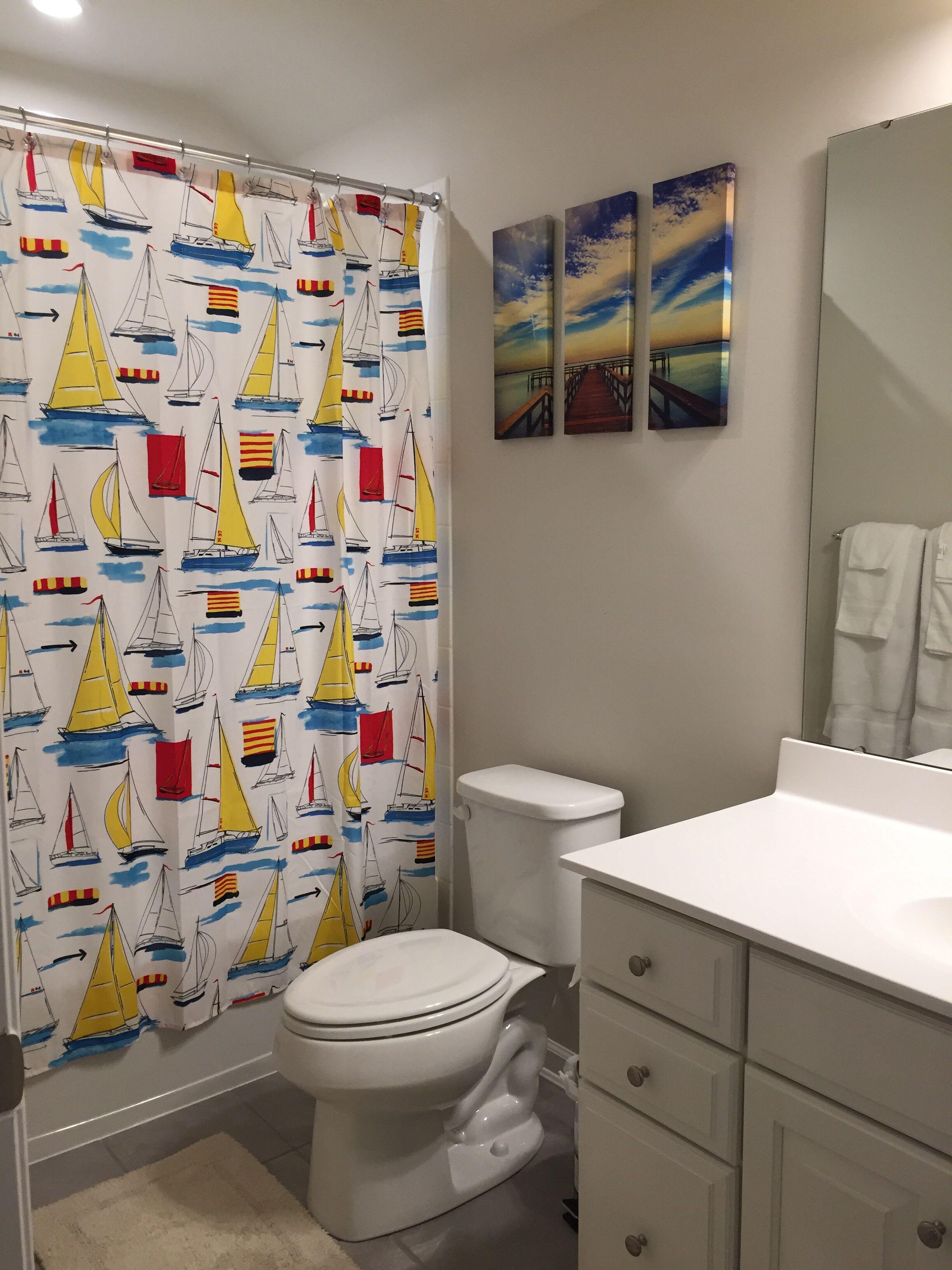Un baño con cortina de ducha con motivos de velero, un inodoro, un lavabo y dos cuadros colgados en la pared.