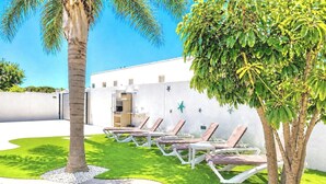 Un área junto a la piscina con sillones de descanso, una palmera y un edificio con fachada blanca.