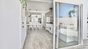 Una cocina moderna con armarios blancos, un comedor con sillas blancas y un ventanal corredero que da a un balcón con mobiliario exterior.