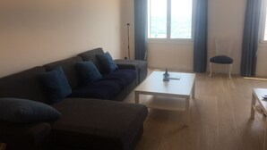 A living room with a dark-colored sofa, a white coffee table, and a chair. There is a window with curtains and a view of the outside. - Port bo's treasure  (Calella de Palafrugell)