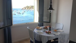 A dining area with a table set for two, a view of boats on the water, and a large window with curtains. - Port bo's treasure  (Calella de Palafrugell)