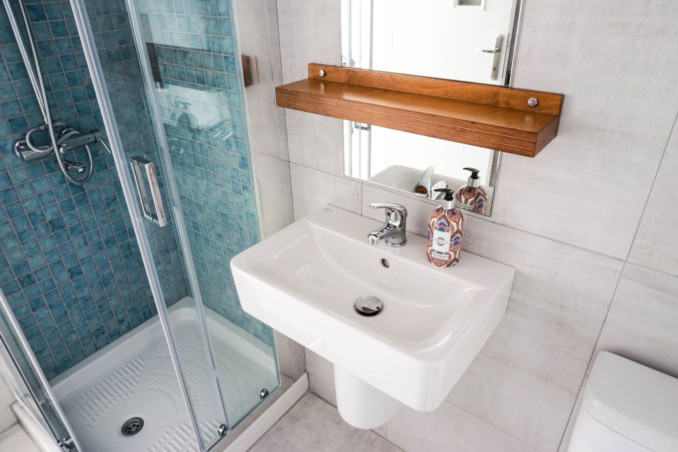 A bathroom with a glass shower enclosure, a white sink, and a wooden shelf above the mirror.