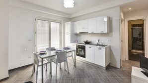 Moderni keittiö, jossa on valkoiset kaapit, ruokapöytä tuoleineen, sisäänrakennettu uuni ja suihkutila, joka on näkyvissä viereisen oven läpi.
