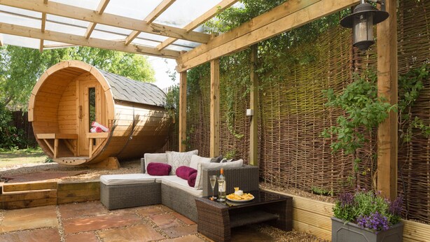 Una struttura a forma di botte in legno con una panca e dei cuscini, un divano in vimini e una zona patio con pavimento in pietra.