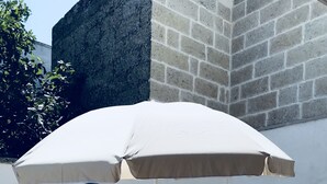 Een terras buiten met een witte parasol, twee gestreepte stoelen en een betegelde vloer.